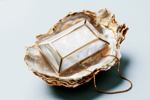 A rectangular resin lantern with gold metal framing rests inside an open oyster shell, with a gold chain draped over the shell against a light gray background.