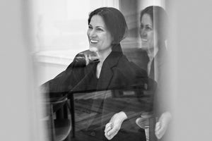 Black and white portrait of a Yliana Yepez in a dark blazer, seen through a reflective glass surface with her reflection visible behind it.